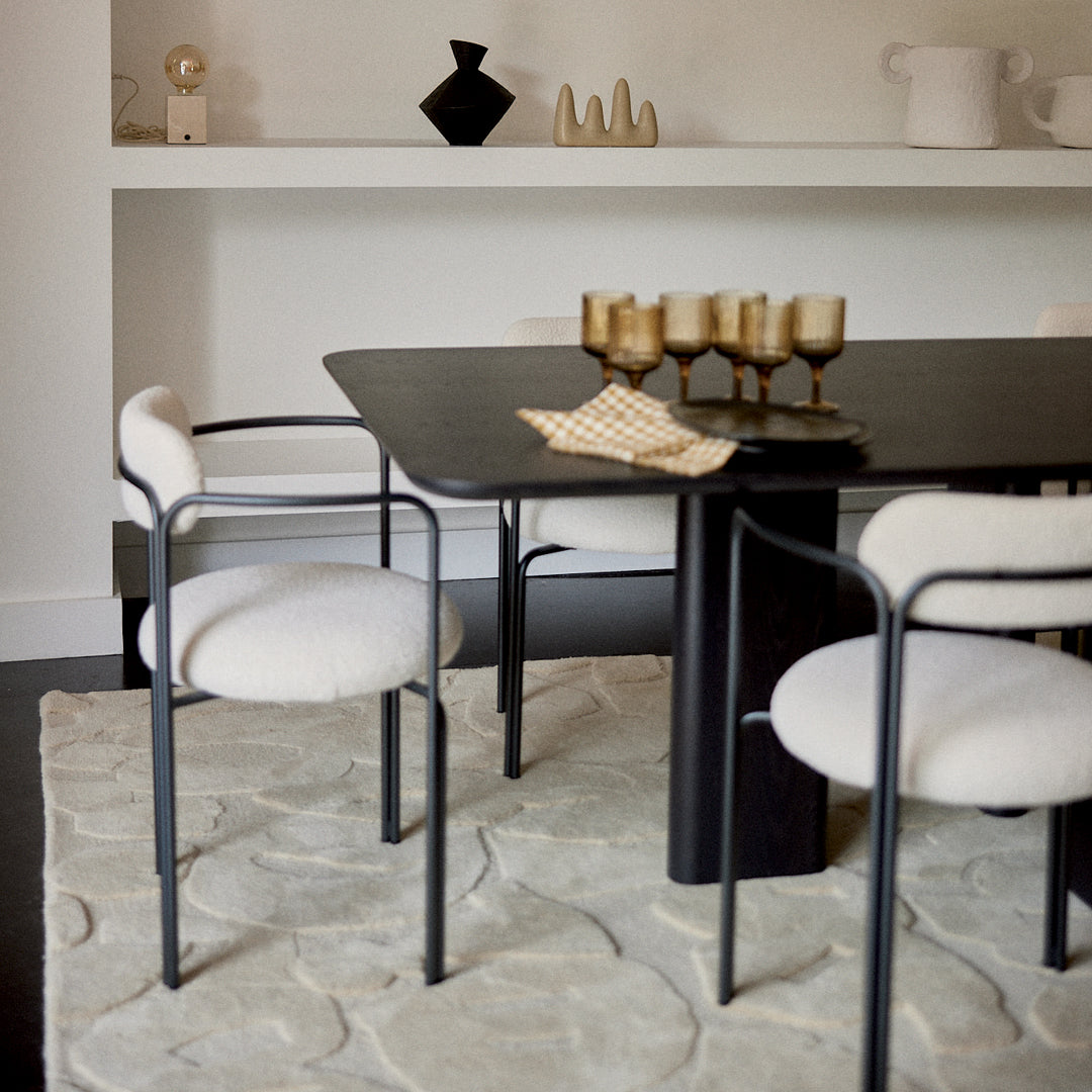 Elegant black table with white upholstered chairs, textured light rug, and warm gold accents. Modern minimalist dining setup.