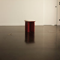Red minimalist stool with a smooth white top, set against a dark floor and neutral wall. Stylish and contemporary design.