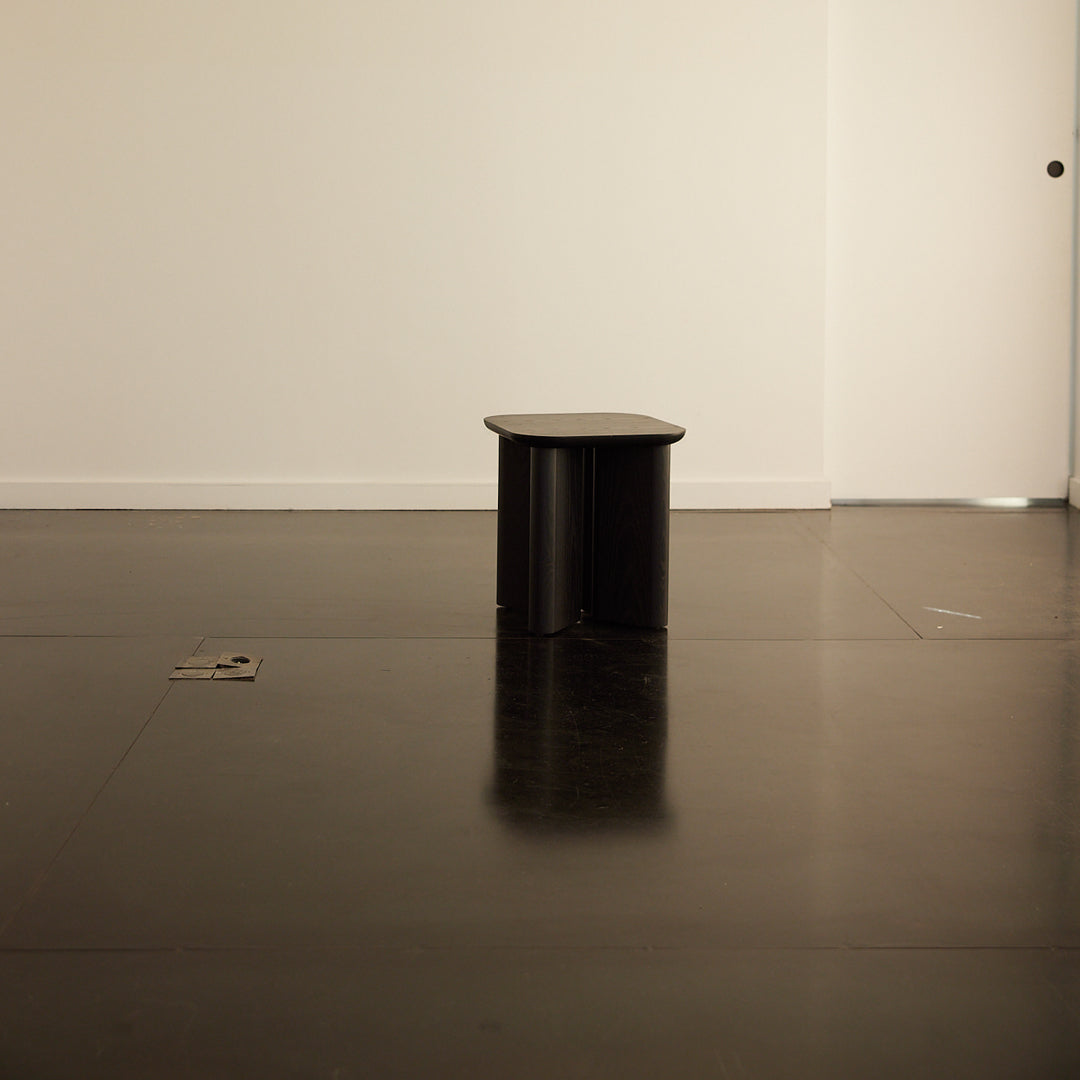 Black minimalist stool on a glossy black floor with subtle reflections and a plain white background.