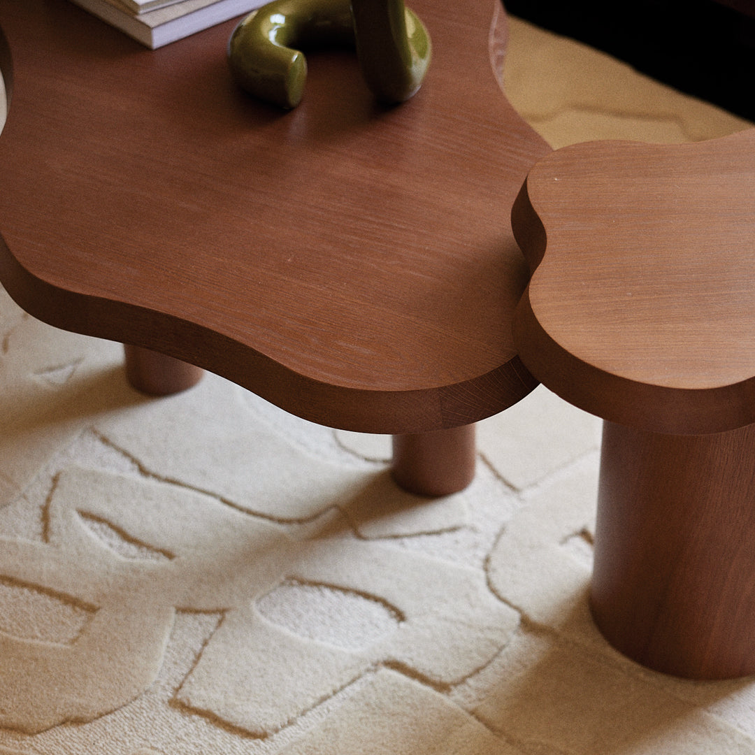Brown wooden coffee table with a unique shape on a textured cream rug, featuring subtle geometric patterns.