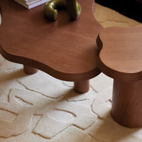 Brown wooden coffee table with a unique shape on a textured cream rug, featuring subtle geometric patterns.