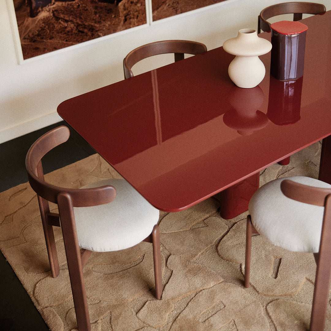 Glossy burgundy dining table with cream upholstered chairs on a textured beige rug. Modern and stylish decor elements.