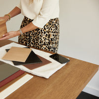 Woman in leopard print skirt arranging wood and colored samples on wooden table with a black phone nearby.