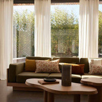 Natural light-filled room with olive green sofa, patterned cushions in mustard, white, and floral accents, wooden coffee table.
