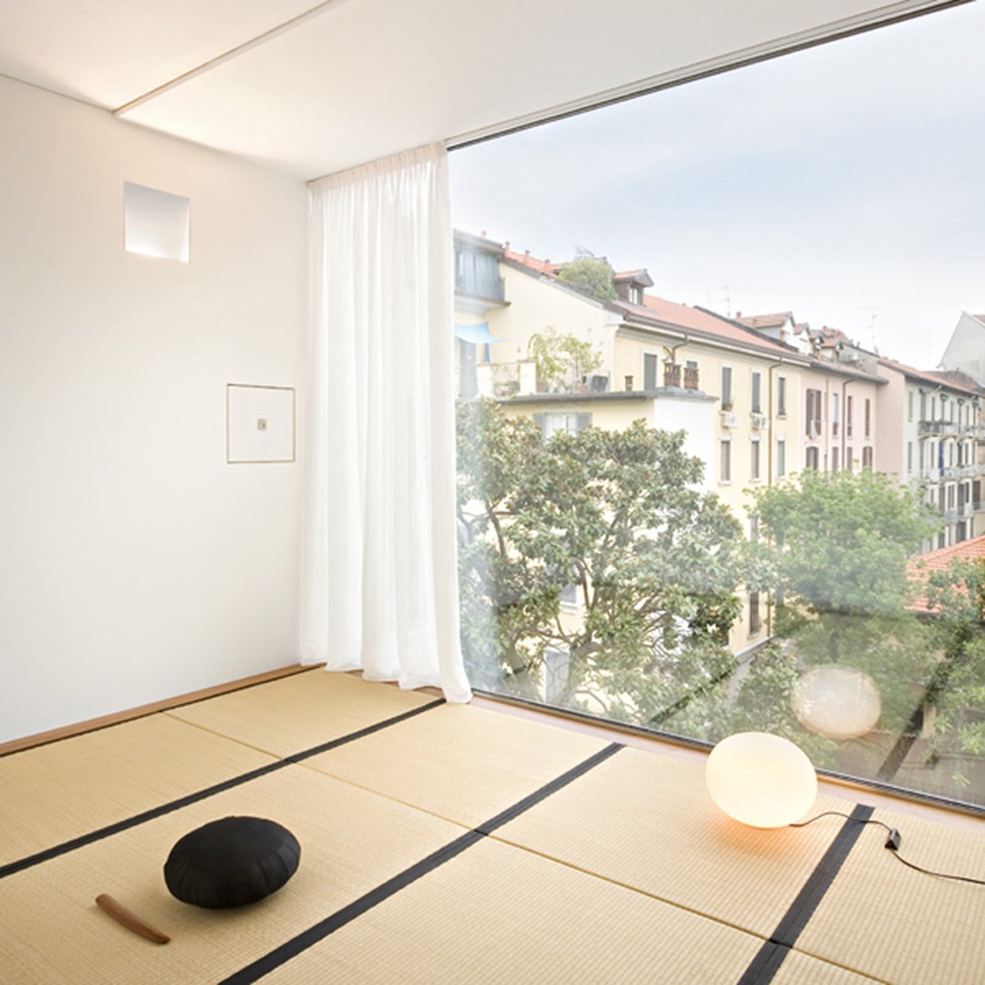 Minimalist room with natural wood tones, white curtains, and a black stone accent. Soft light from a round lamp.