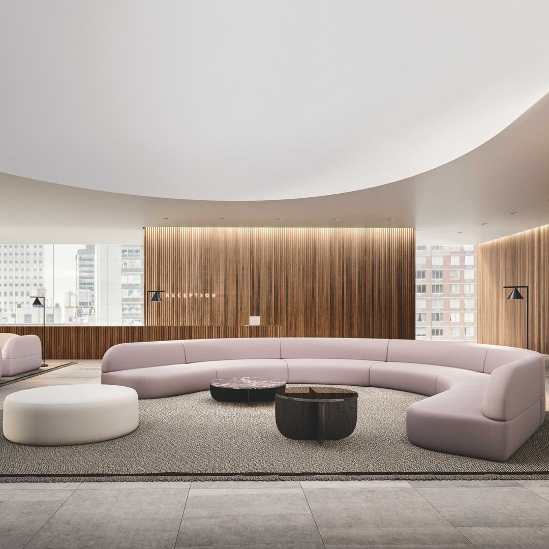 Curved soft pink sofa with white accent and dark round coffee table, set against a textured gray carpet and warm wood paneling.