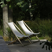 Two minimalist beige canvas loungers with natural wood frames, set against a lush green backdrop.