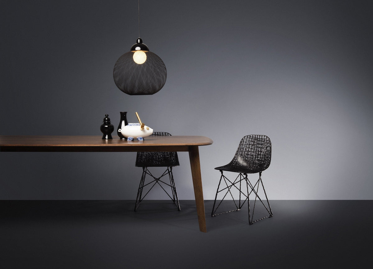 Black woven pendant light above a wooden table; black chairs with intricate patterns; muted gray backdrop.