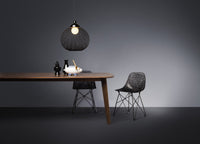 Black woven pendant light above a wooden table; black chairs with intricate patterns; muted gray backdrop.