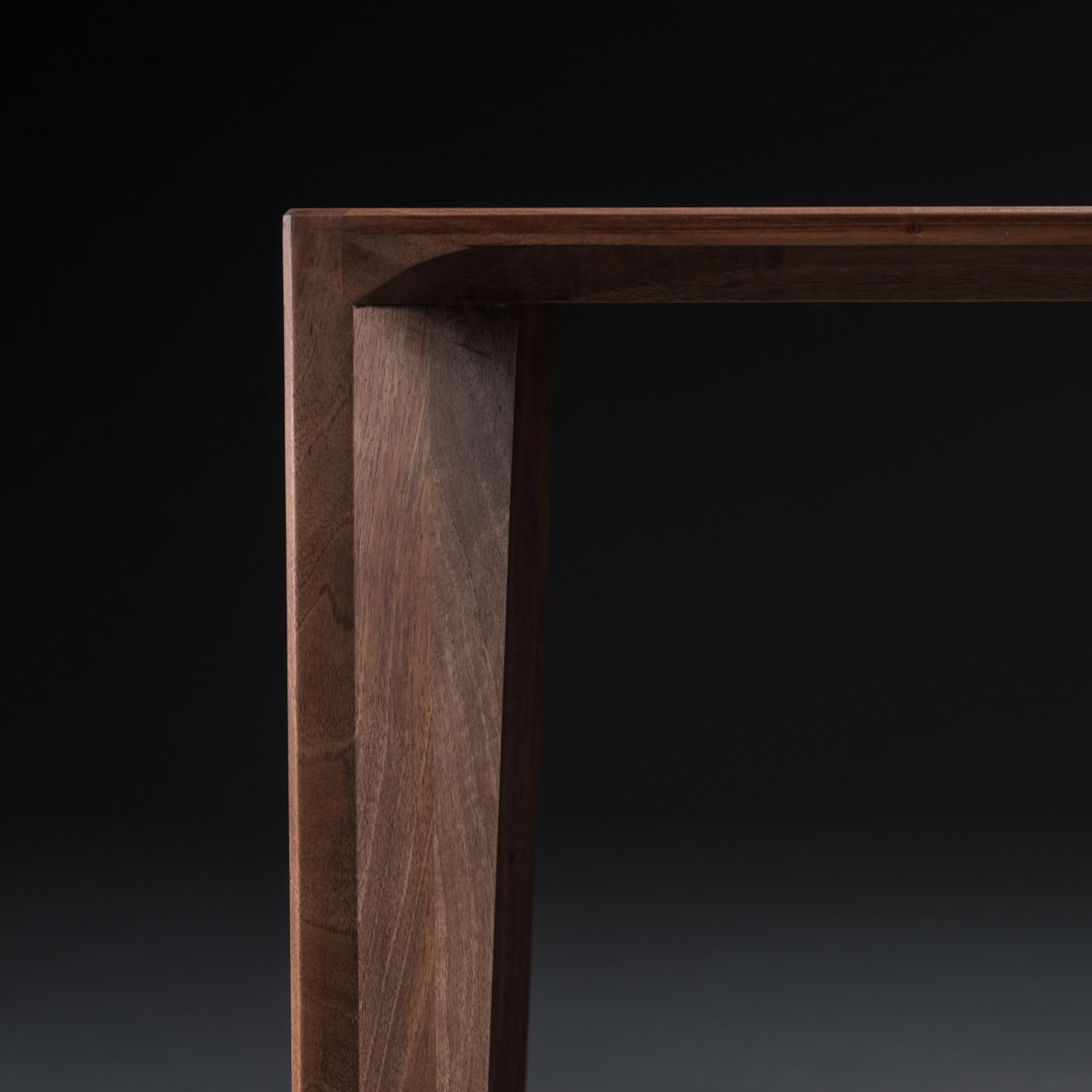 Wooden table corner in rich walnut finish, showcasing smooth texture and elegant angular lines against a dark background.