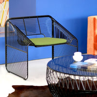Black wireframe chair with a green cushion, paired with a geometric glass table against a blue backdrop.