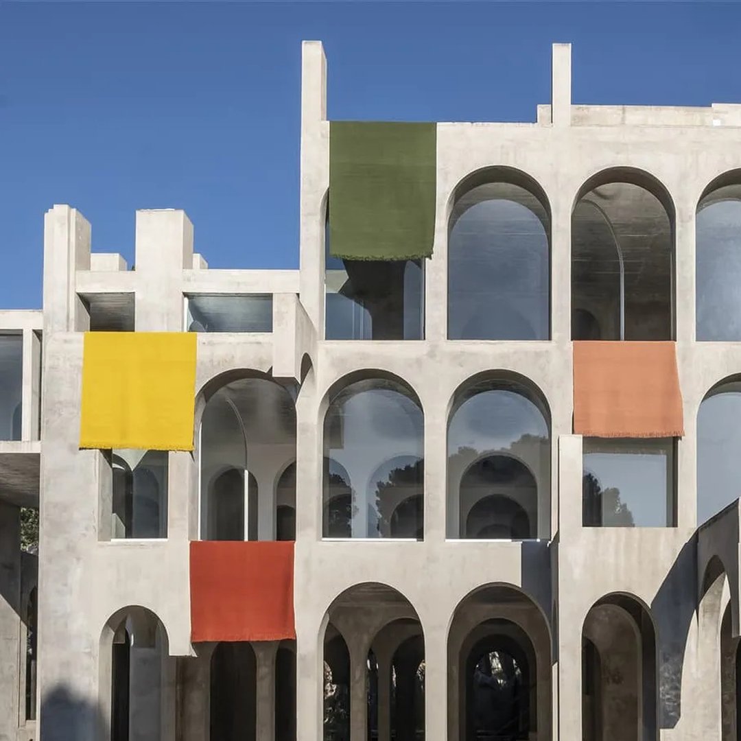 Colorful fabrics in yellow, green, and orange drape over a modern concrete structure against a clear blue sky.
