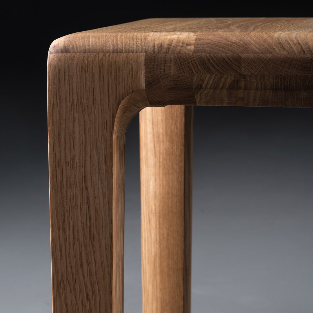 Close-up of a wooden table corner featuring warm, rich brown tones and smooth, rounded edges. Natural grain pattern visible.
