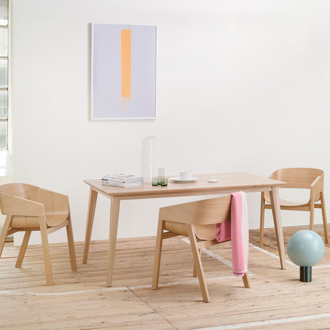 Light wood dining table with minimalist design, featuring beige chairs and a pastel pink throw against a white backdrop.