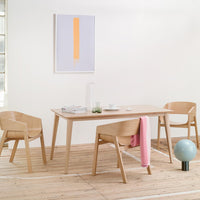 Light wood dining table with minimalist design, featuring beige chairs and a pastel pink throw against a white backdrop.
