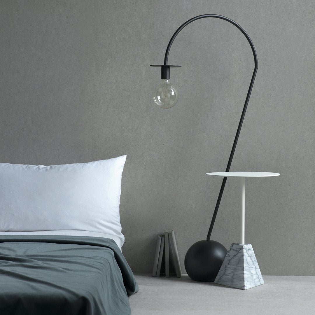 Minimalist black floor lamp with clear globe, gray bedding, white pillow, and geometric marble side table.