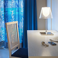 Blue and white patterned curtains complement a sleek, modern desk lamp in a bright, airy workspace.