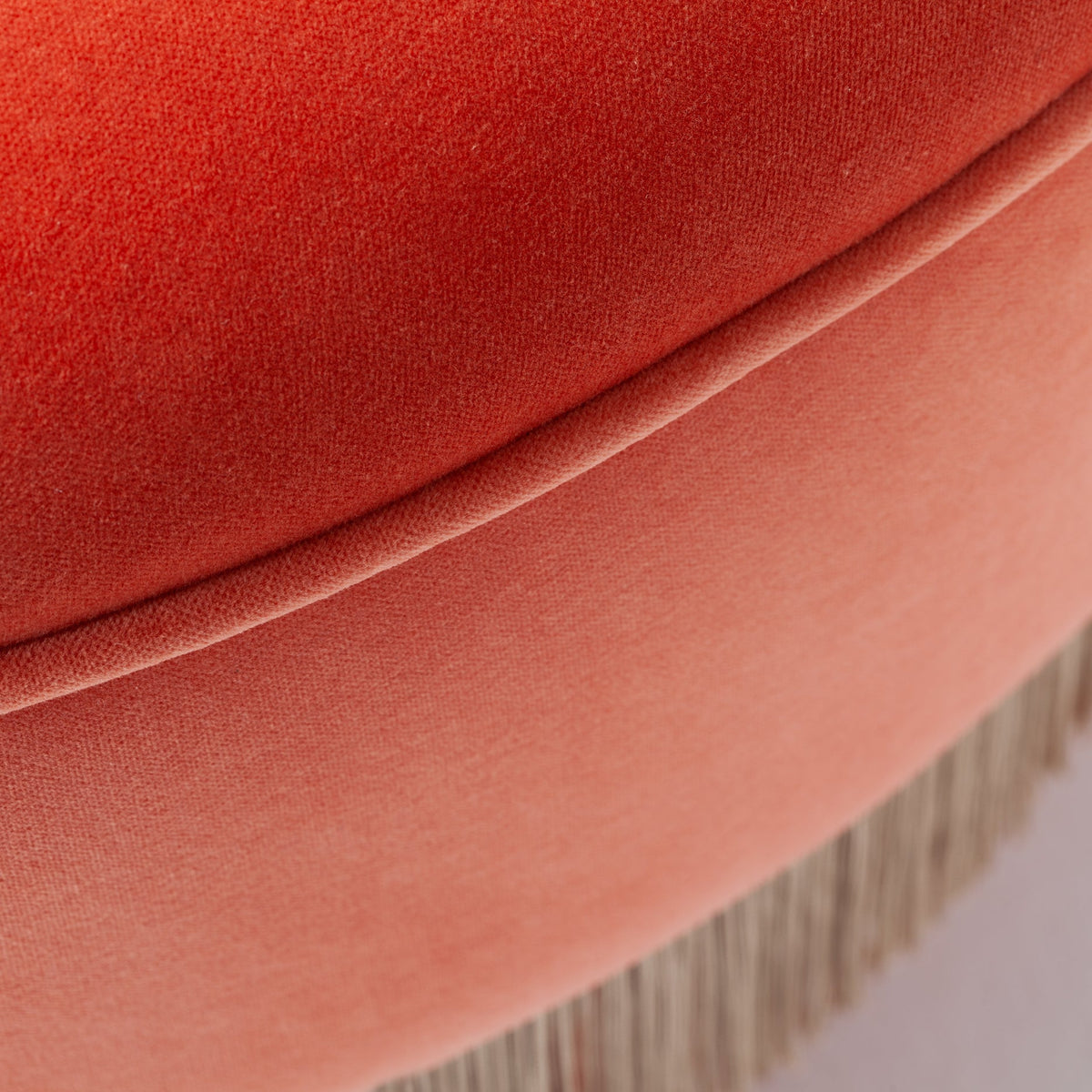 Coral velvet ottoman featuring a smooth gradient and delicate fringe detail. Perfect for a stylish accent.