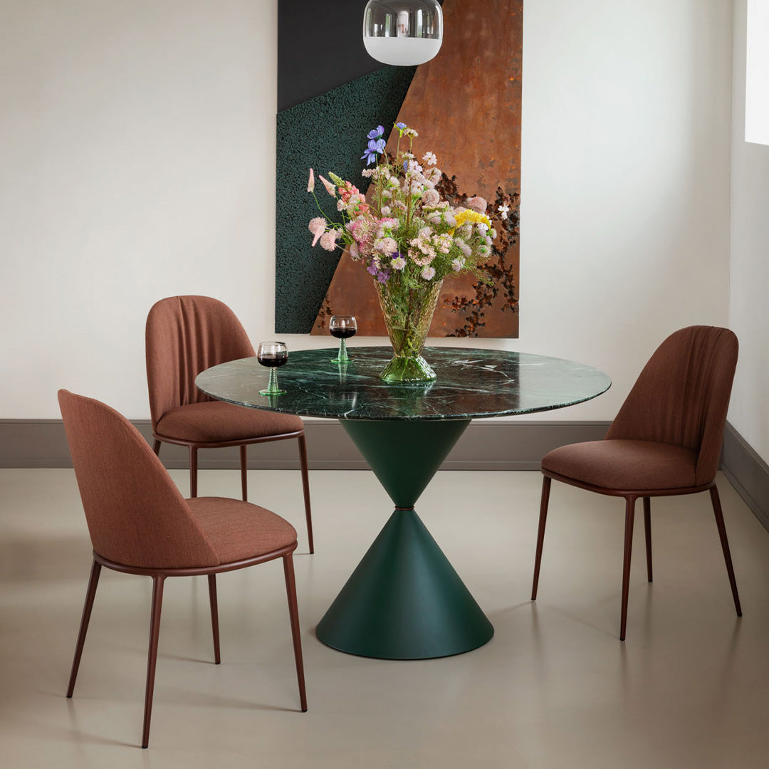 Modern round glass dining table with a green geometric base, surrounded by rust-colored upholstered chairs. Soft floral centerpiece.