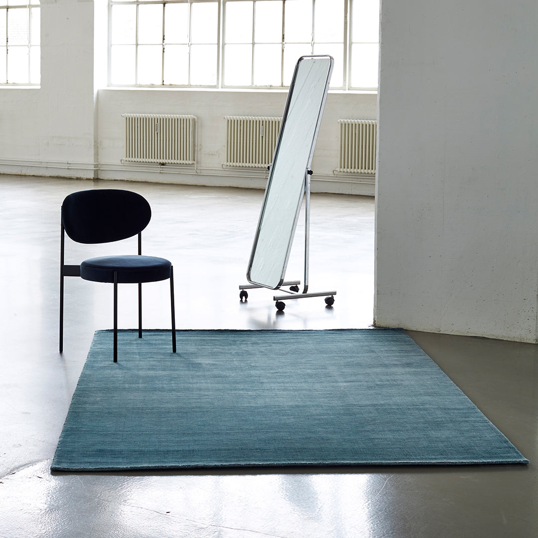 Teal rectangular rug with a smooth texture, paired with a sleek black chair and modern mirror in a minimalist space.