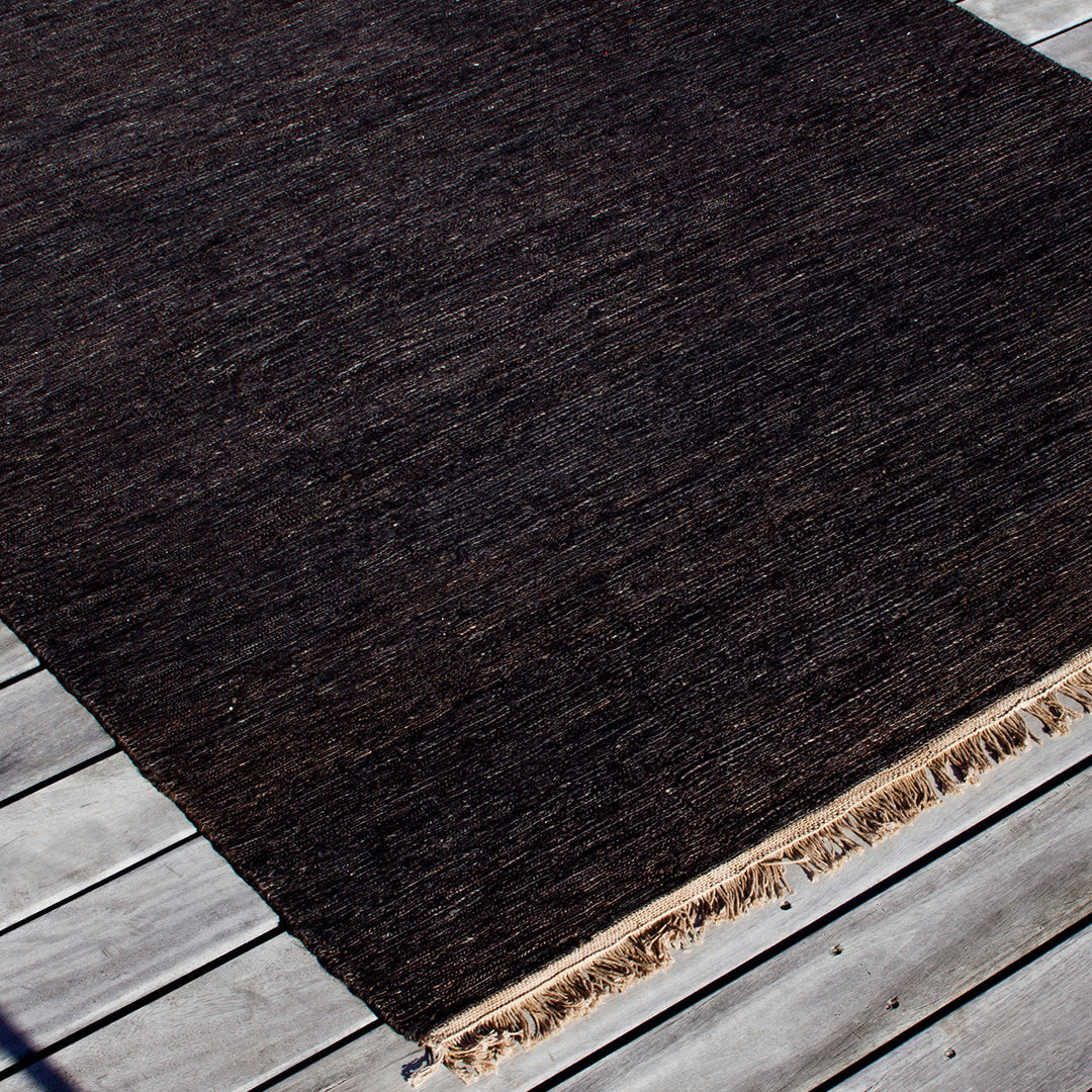 Dark brown textured rug with subtle variations, featuring fringed edges on a light wooden surface.