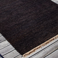 Dark brown textured rug with subtle variations, featuring fringed edges on a light wooden surface.