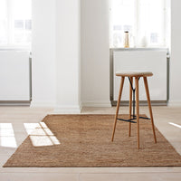 Natural brown jute rug with a subtle texture, paired with a minimalist wooden stool in a bright, airy room.