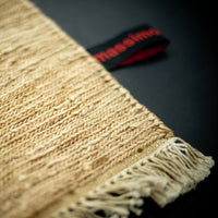 Textured woven mat in natural beige tones, featuring intricate patterns and fringe edges, with a bold red and black label.