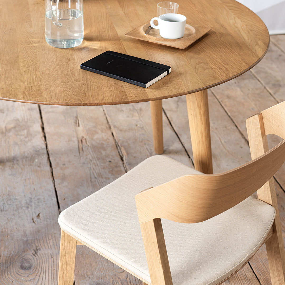 Light wood dining table with a round top, paired with a cream-colored chair and a black notebook on the surface.