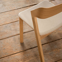 Natural wood chair with light beige upholstery against rustic wooden flooring. Simple, modern design and earthy tones.