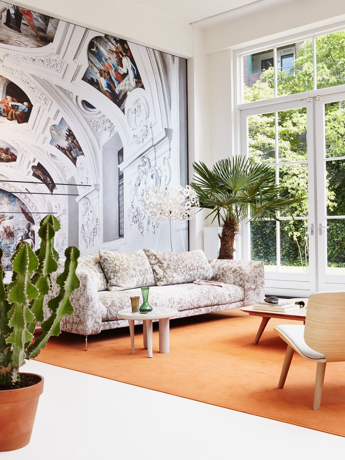 "Light floral-patterned sofa on an orange rug, surrounded by greenery and a decorative wall mural."