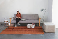 Gray sofa with wooden arms, orange area rug, and textured cream ottoman against a minimalist background.