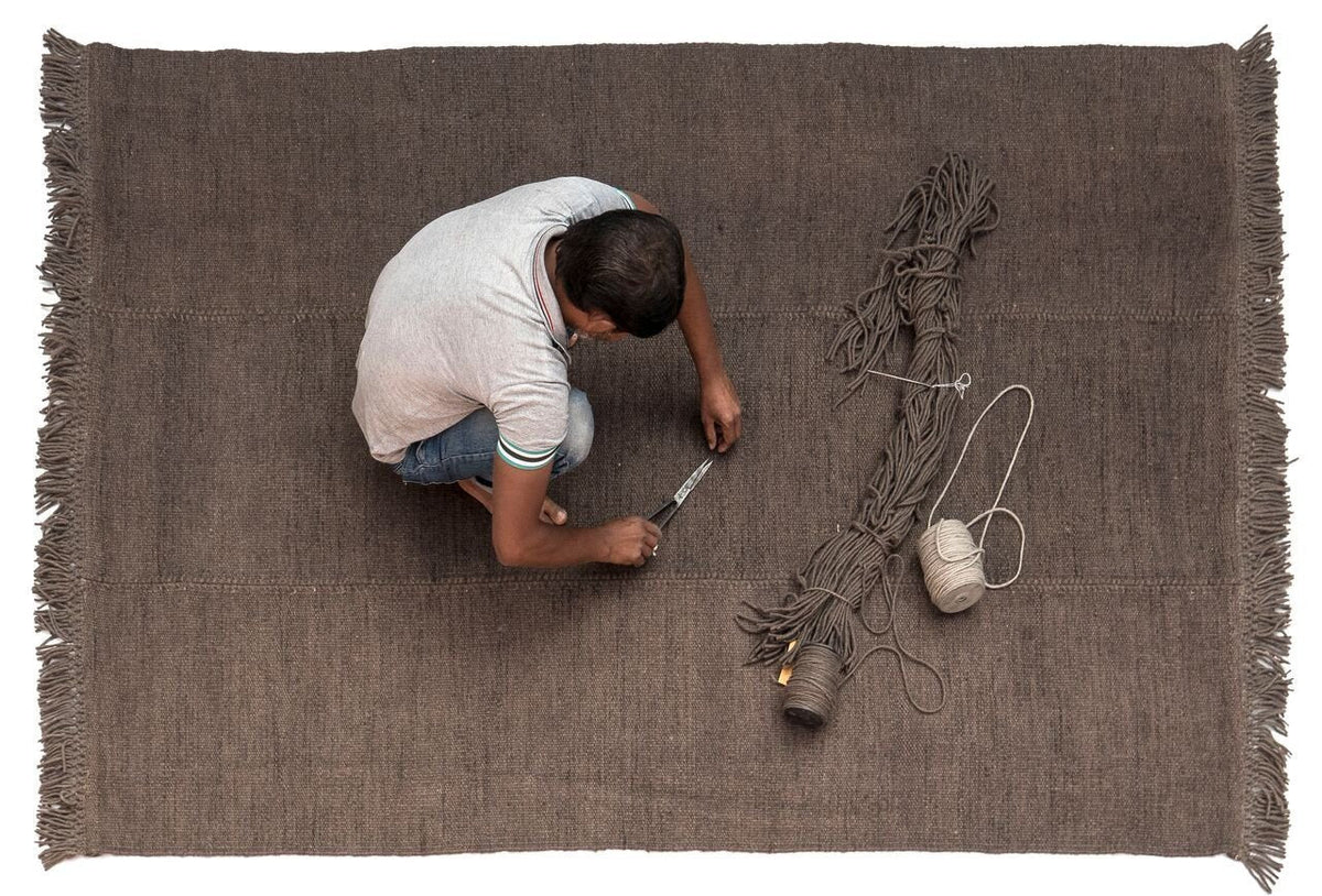 Brown woven rug with a textured pattern and fringed edges, featuring a neutral color palette.