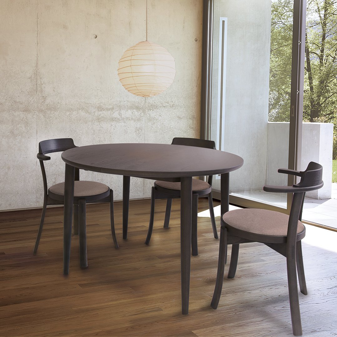Round brown dining table with matching brown chairs, set against a light gray wall and warm wood flooring.