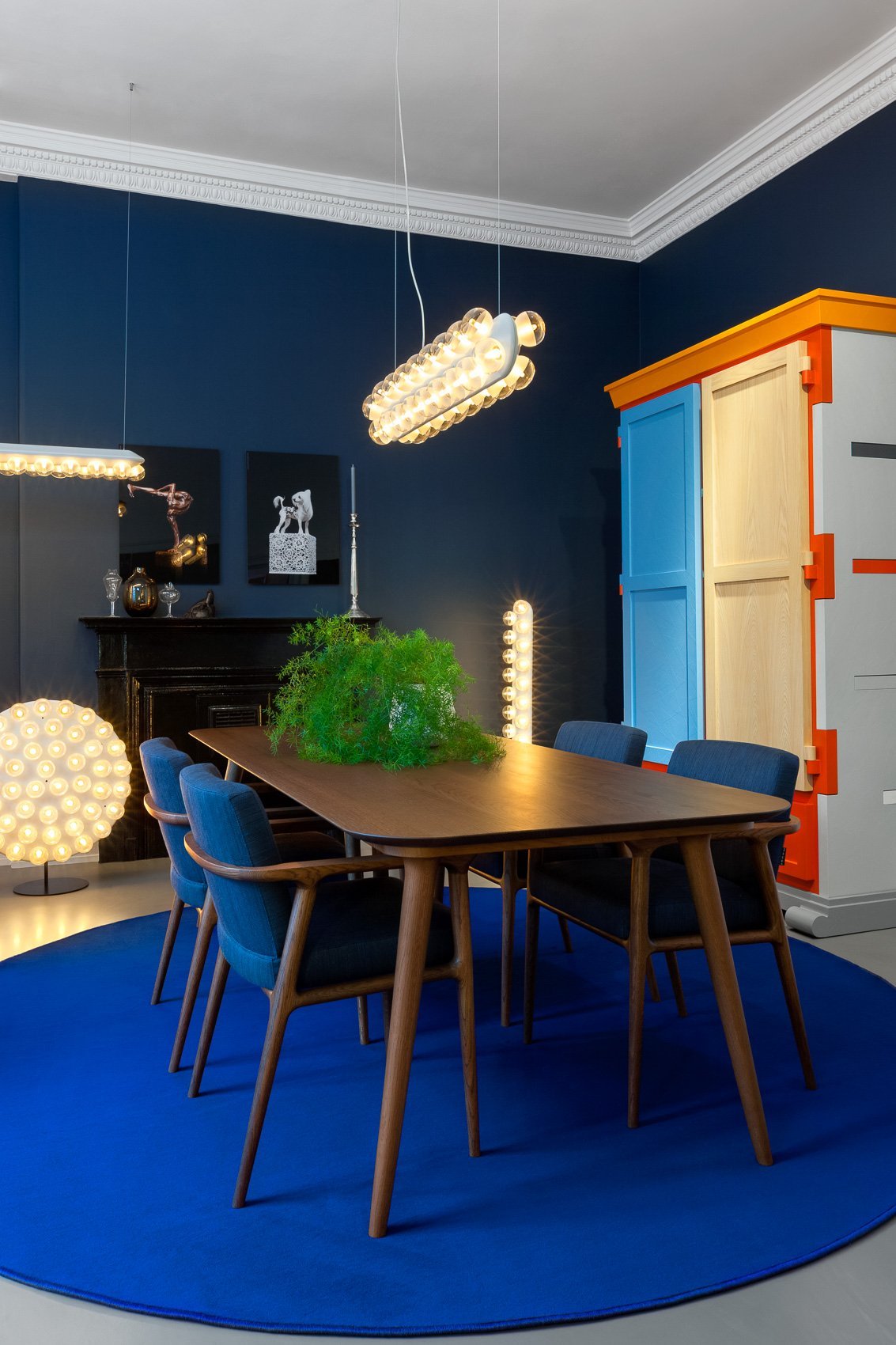 Modern dining room featuring a blue round rug, walnut table, and stylish blue chairs against a navy backdrop.