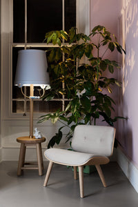 Stylish light gray lounge chair with natural wood legs, paired with a soft blue lamp and green leafy plant against a pastel pink wall.