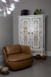 Colorful floral patterns on a white cabinet, complemented by a brown armchair and neutral gray walls.