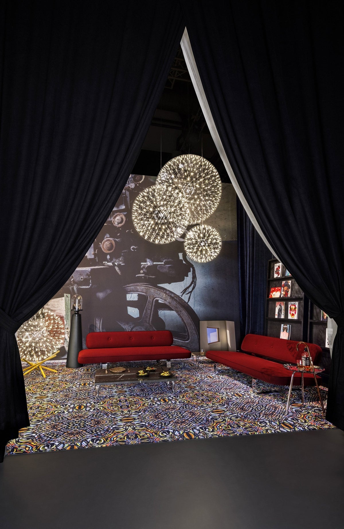 Red sofas contrast with a multicolored patterned rug, framed by dark blue curtains and illuminated by pendant lights.