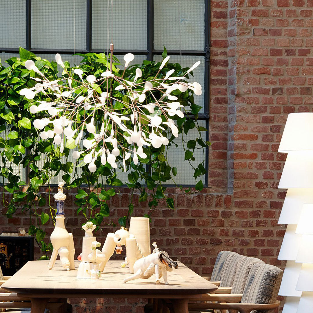 "Colorful modern chandelier with white leaf-like accents, set against a brick wall and green foliage."