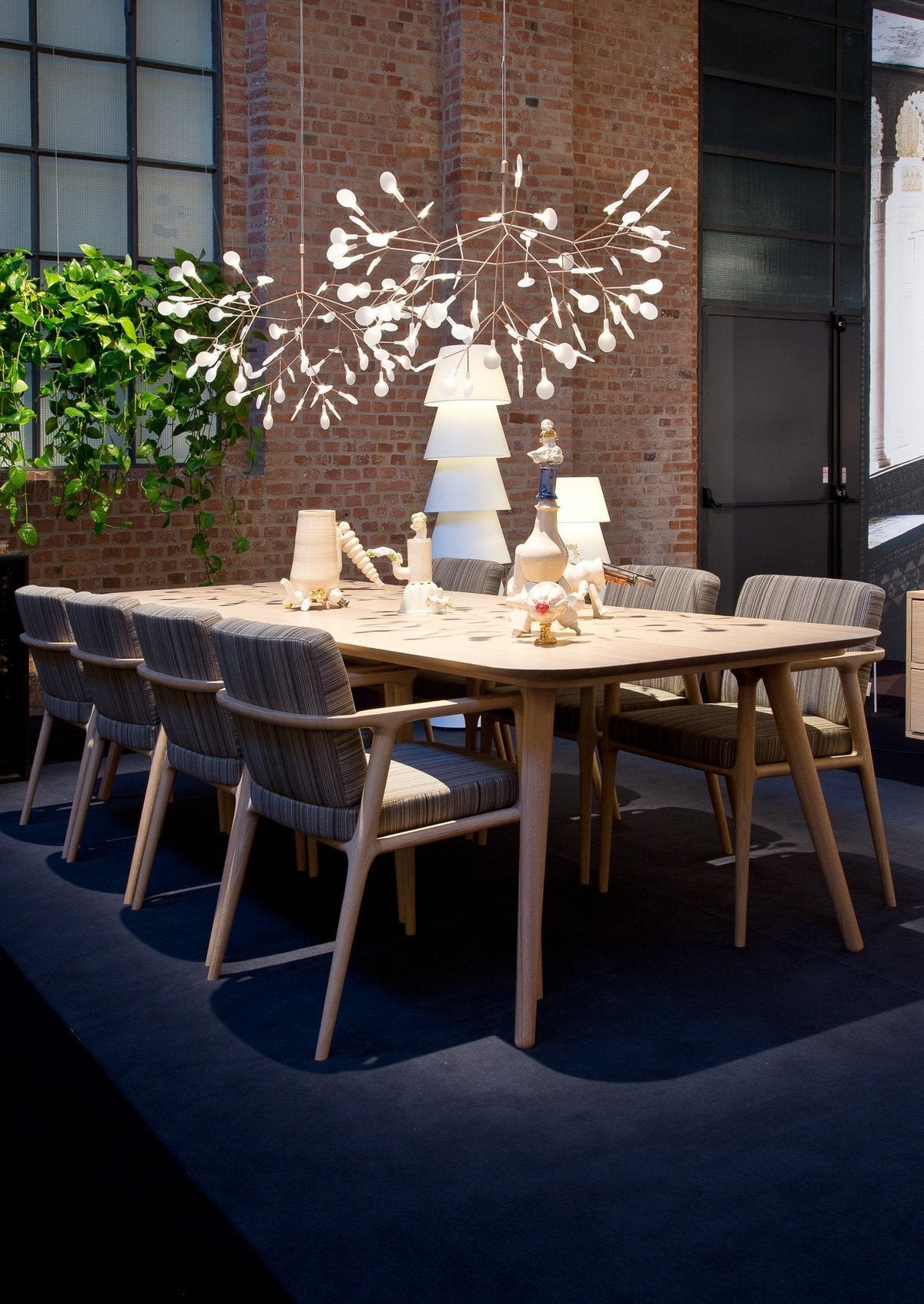 Light wood dining table with gray upholstered chairs, decorative white hanging lights, and green foliage accents.