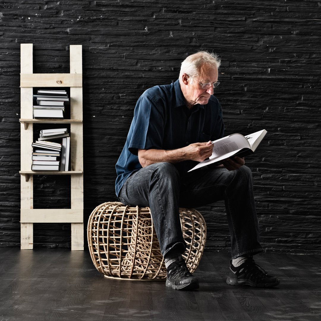 "Natural woven rattan pouf in neutral tones, set against a dark textured wall and minimalist white bookshelf."