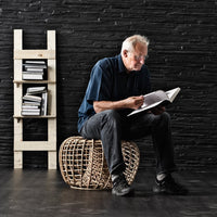 "Natural woven rattan pouf in neutral tones, set against a dark textured wall and minimalist white bookshelf."