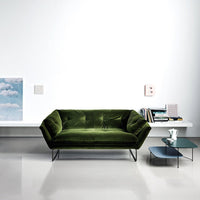 Green velvet sofa with a tufted design, set against a minimalist white wall and sleek glass side table.
