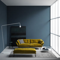 Olive green velvet sofa and ottoman against deep blue wall, complemented by a gray rug and modern floor lamp.