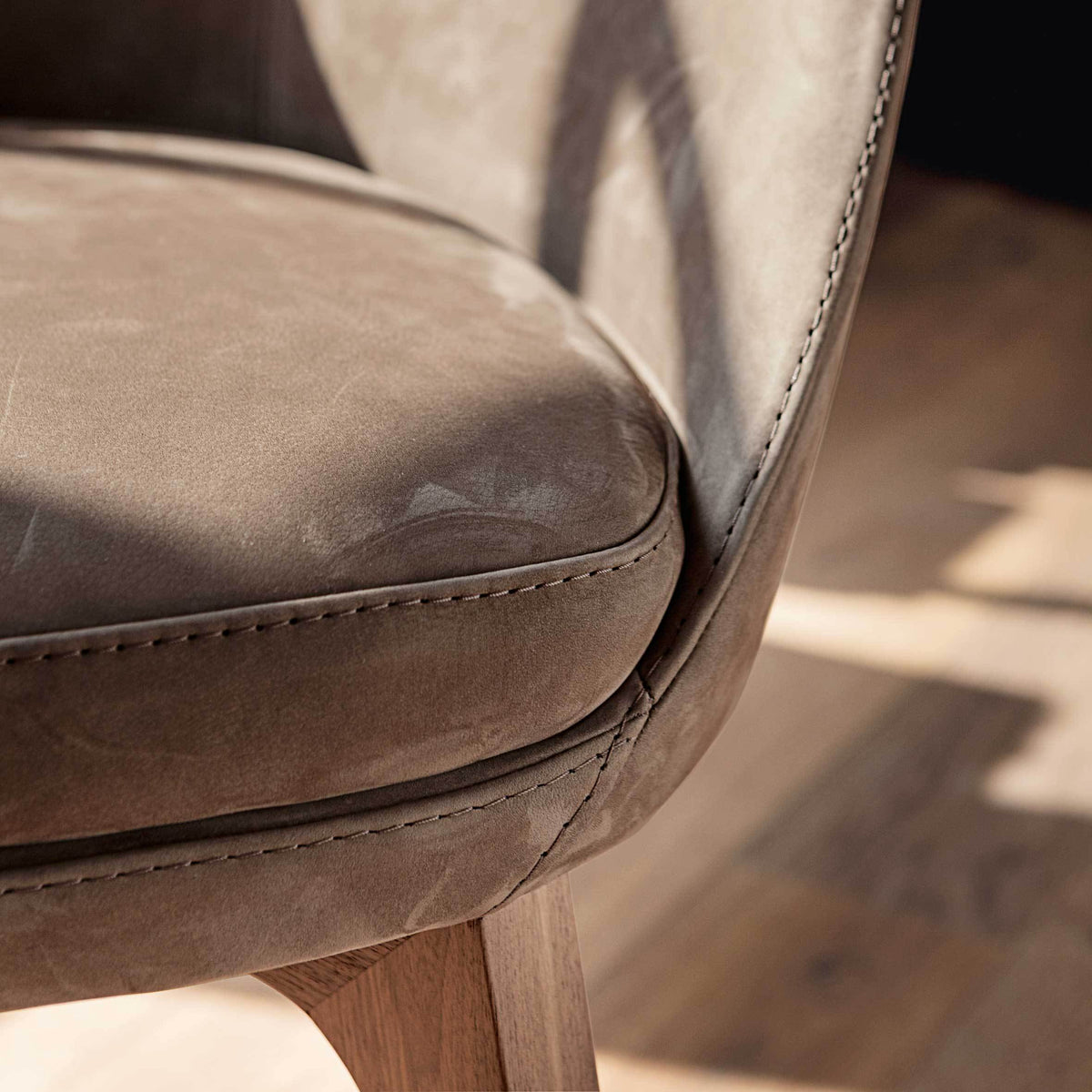 Taupe leather chair with subtle texture and smooth stitching, resting on a natural wood floor backdrop.