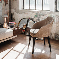 "Soft beige upholstered chair with sleek black legs, set against a textured wall and warm wood flooring."
