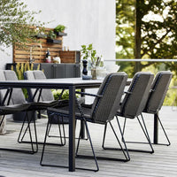 Black woven chairs and a sleek black table on a wooden deck, surrounded by greenery. Modern outdoor dining set.