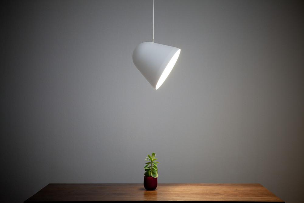 Modern white pendant lamp illuminating a small green plant on a wooden table against a gray background.