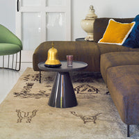 Black cone table with a circular top, set on a beige patterned rug, complemented by green and yellow accent pillows.