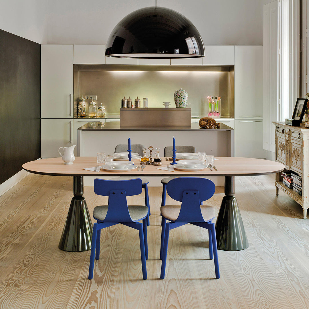 Oval light wood dining table with black and blue accent chairs, set against a modern kitchen backdrop.